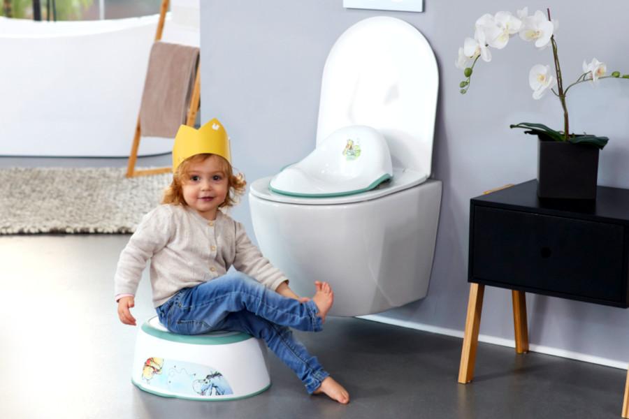 Ein kleines Mädchen mit Krone auf dem Kopf sitz barfüßig auf dem Tritthocker "kasimir" vor der Toilette.