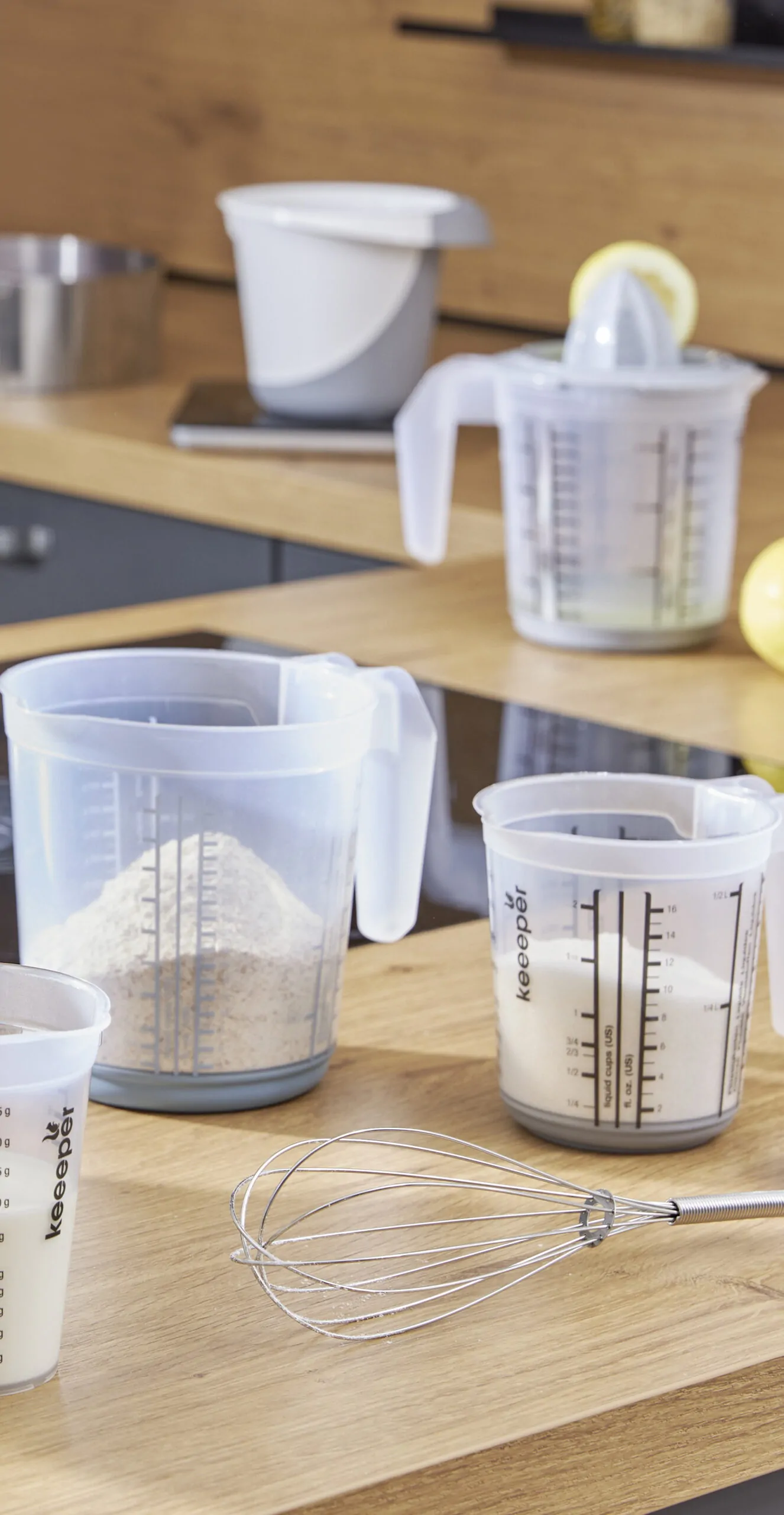 Transparent measuring jugs ''matteo'' and cups with measurement markings on a kitchen counter, alongside baking ingredients and a whisk.