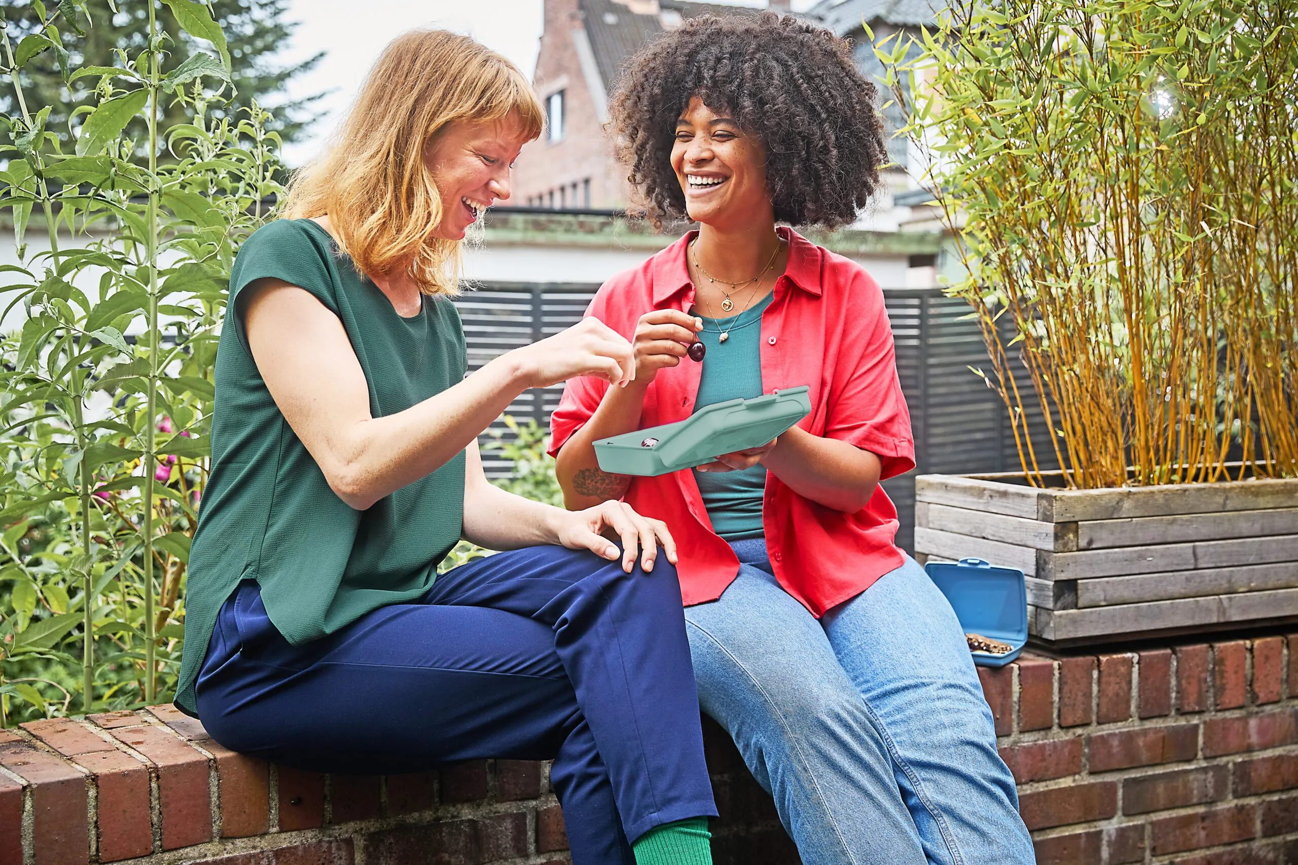 Two women sitting outdoors in a garden, sharing berries from a reusable food container ''luca'' and smiling together.