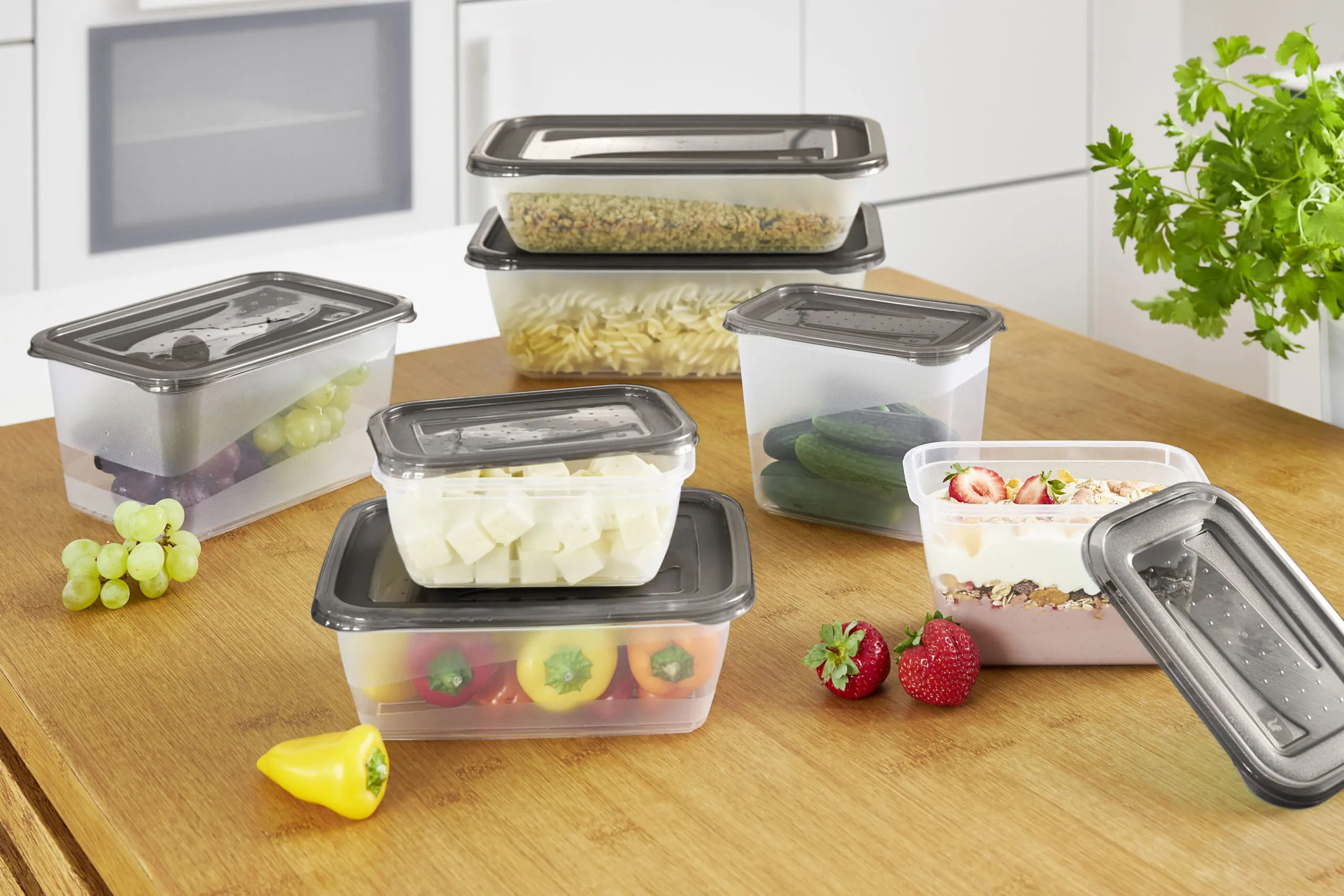 Food storage containers ''fredo fresh'' with dark lids arranged on a wooden table, filled with fresh fruit, vegetables, pasta, and prepared meals.
