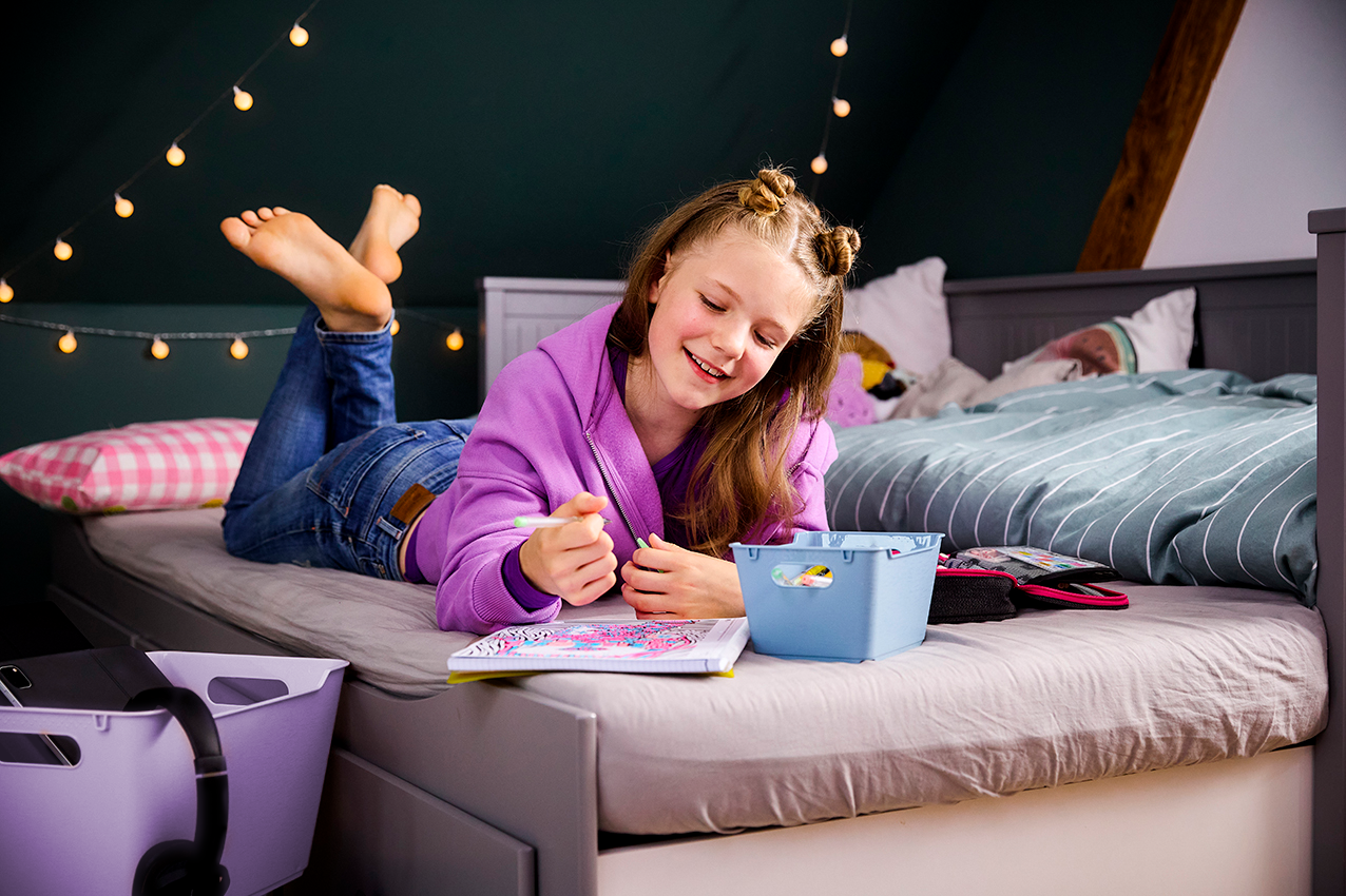 Ein Mädchen malt entspannt auf dem Bett und nutzt dabei eine kleine blaue Aufbewahrungsbox für Stifte und Bastelmaterial.