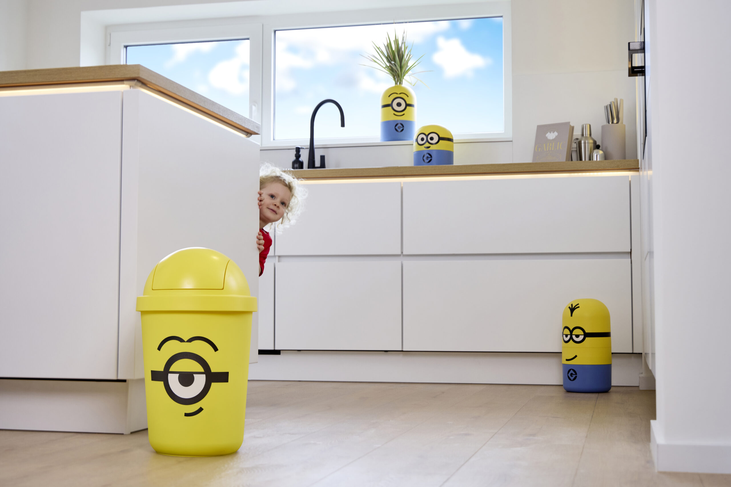 Modernes Zuhause mit gelben Minions-Abfallbehältern in der Küche, während ein Kind neugierig hinter der Kücheninsel hervorschaut.
