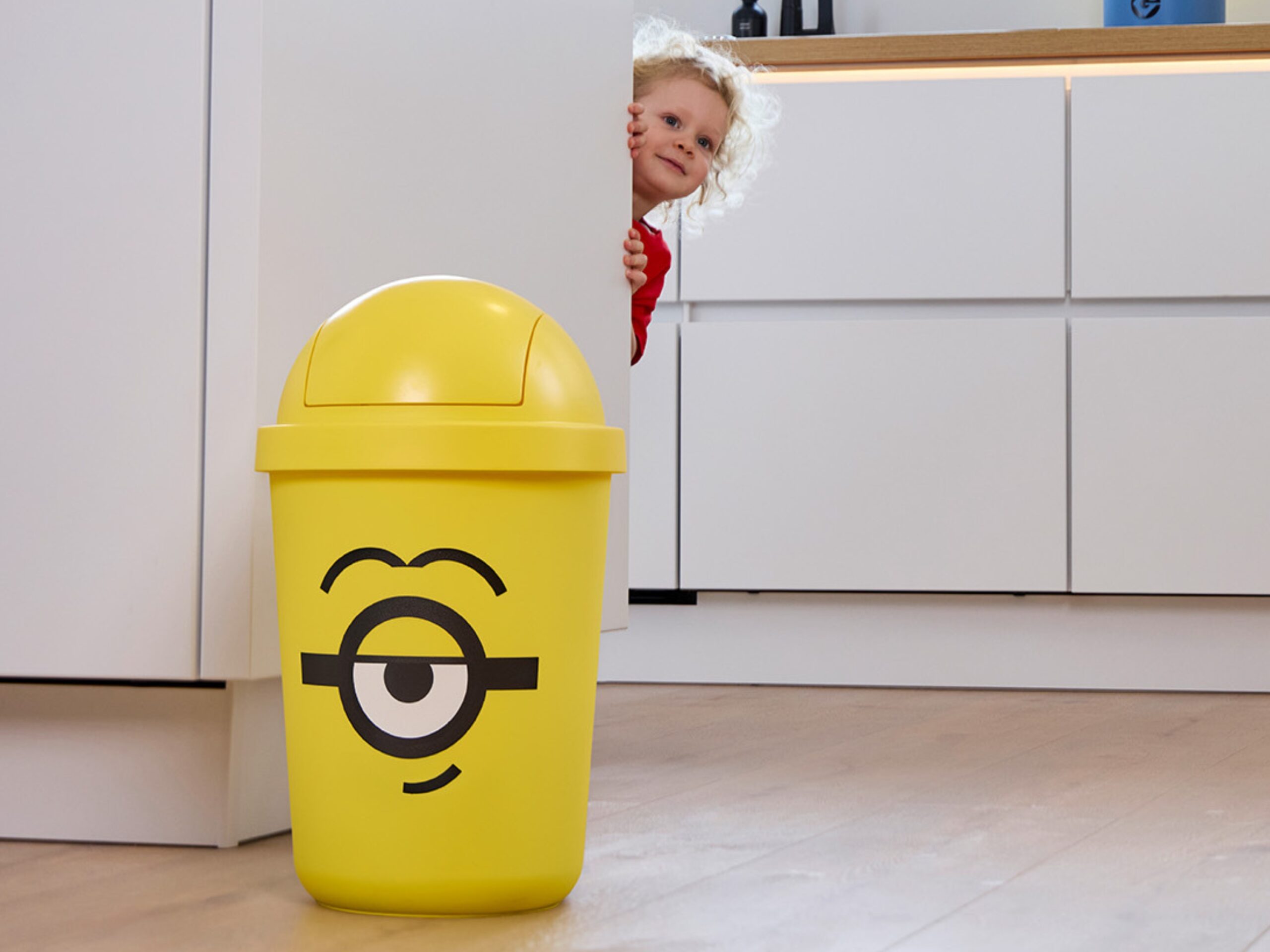 A yellow Minions-themed bin stands on the floor while a child peeks from behind a cabinet in the background.